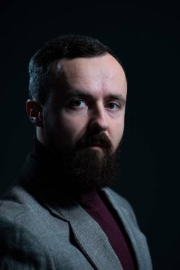 Ceket ve süveter giymiş sakallı bir milenyum erkeğinin dramatik portresi. Karanlık bir arka planda.