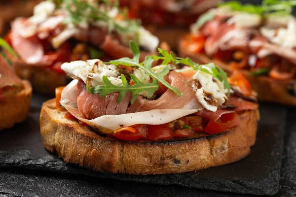 stock image Parma ham and tomato bruschetta served on black slate with mozzarella, wild rocket leaves and balsamic vinegar.