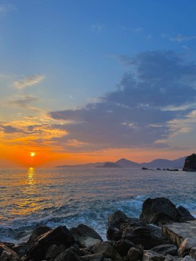 Gün batımında Rocky Sahili. Dalgaların yavaşça kıyıya vurduğu manzaralı bir kayalık kıyı şeridi. Akşam güneşi açık denizde ve engebeli kayalıklarda ılık altın renkler bırakır..