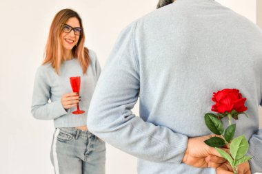 A man holding a red rose behind his back while his girlfriend is standing on background and smiling. 