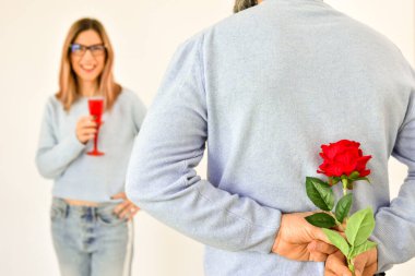 A man holding a red rose behind his back while his girlfriend is standing on background and smiling. 