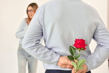 A man holding a red rose behind his back while his girlfriend is standing on background and smiling. 