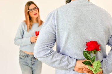 A man holding a red rose behind his back while his girlfriend is standing on background and smiling. 