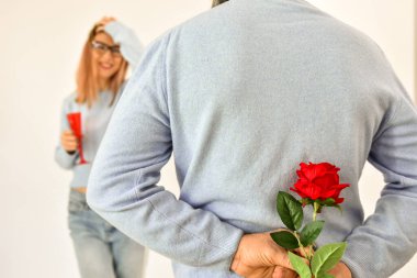 A man holding a red rose behind his back while his girlfriend is standing on background and smiling. 