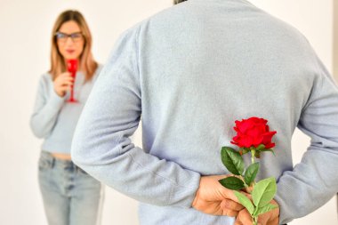 A man holding a red rose behind his back while his girlfriend is standing on background and smiling. 