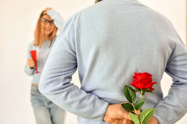 A man holding a red rose behind his back while his girlfriend is standing on background and smiling. 