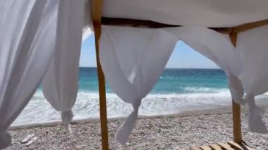 Beyaz kumsal gölgeleri. Sahilde, deniz kenarında bir çardak. Rocky plajı. Perdeler rüzgarda gelişir. Gün ışığı yazı. Arnavutluk 'un güneyinde, Himare' deki Livadi Sahili 'nde. yatay video, HD 