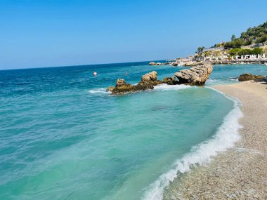 Rocky beach and crystal turquoise water of Ionian Sea in Albania. Calm and relaxing view with flowers. summer holidays concept. Copy space. clipart