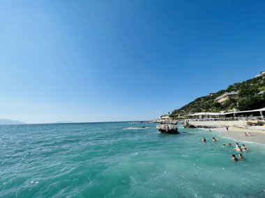 Rocky beach and crystal turquoise water of Ionian Sea in Albania. Calm and relaxing view with flowers. summer holidays concept. Copy space. clipart