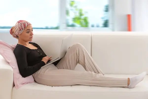 头戴围巾的女人躺在沙发上 膝上型电脑 图库照片