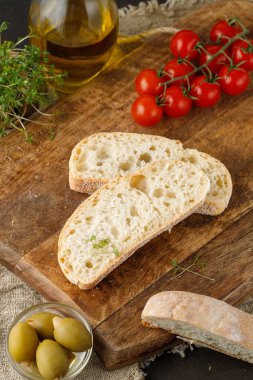Italian ciabatta wheat bread slices, herbs, cherry tomatoes and olives on wooden board over linen towel. Fresh baked sliced ciabatta closeup.