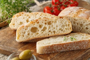 Sliced italian ciabatta wheat bread , herbs, cherry tomatoes and olives on wooden board over linen towel. Fresh baked ciabatta slices closeup.