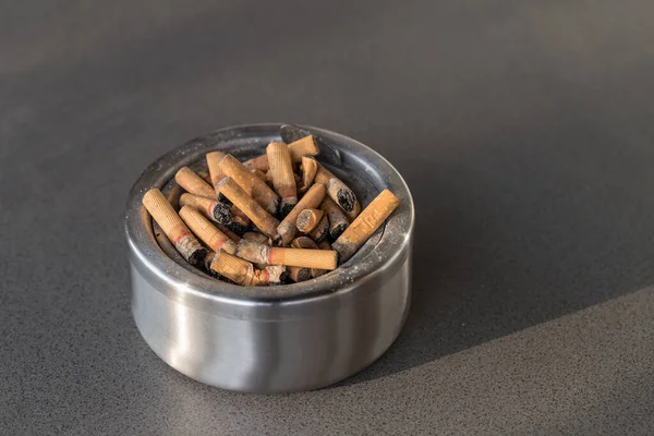 用过的烟头堆在灰色花岗岩石桌上的肮脏金属烟灰缸 很多烟蒂都是尼古丁成瘾的细节 戒烟概念 — 图库照片