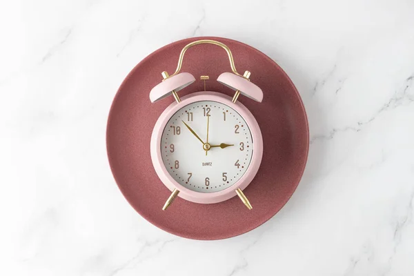 Pink alarm clock on retro red plate on white marble table background. Time to food. Meal on time as element of healthy diet.