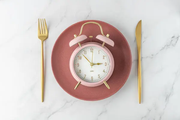 Pink alarm clock on retro red plate with golden fork and knife on white marble table  background. Time to food. Meal on time as element of healthy diet.