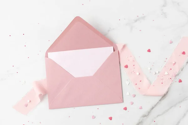 Blank paper card and open retro pink envelope with ribbon and heart shape sprinkles on white marble background. Mockup birthday or valentines day greeting card with copy space.