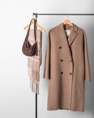 Kahverengi yün çift göğüslü ceket ya da ceket, bej alpaka eşarp ve açık gri duvar zeminine asılı koyu süet çanta. Sıcak yün giysiler. Moda monokrom moda.