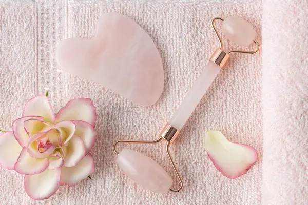 Gül kuvars yüz silindiri ve yaşlanma önleyici cilt masajı için guasha taşı açık pembe havluyla beyaz çiçekler. Yüz masajı için kozmetik araç.