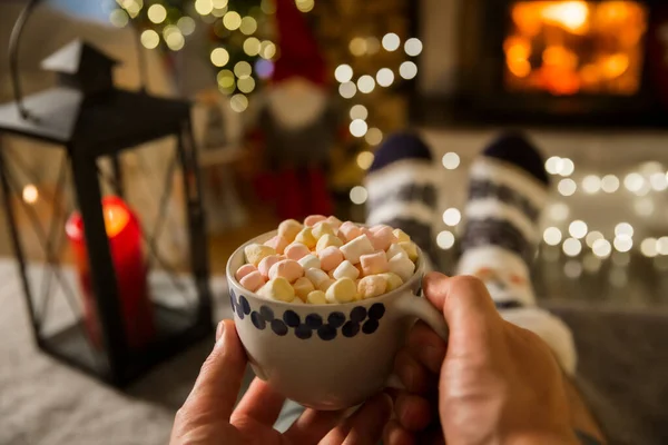 Man enjoying cocoa with marshmallows by warm fireplace and warming up their feet in woolen socks with Christmas ornaments and Christmas tree and decorations background. Cozy Winter, Christmas and