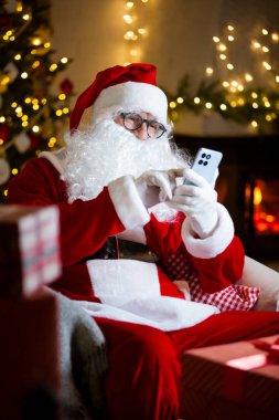 Jolly Santa Claus with eyeglasses scrolls his smartphone, reads heartfelt letters by a cozy fireplace in a cozy festive room with Christmas tree, decorated by twinkling lights. Coming holiday vibes clipart