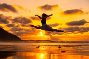 Gün batımında plajda genç bir dansçı arka planda deniz ile bir atlayış yapıyor, güneş suya yansıyor.