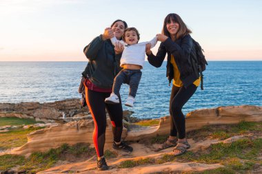 Anne ve çocuğun teyzesi gün batımında deniz kenarında onunla oynuyor.