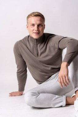 Beyaz arka planda kahverengi süveterli sarışın beyaz model, gülümseyen kameraya bakıyor.