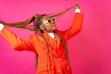 Pembe arka planda örgü ören Afrikalı genç bir kadın, kırmızı elbiseli, eğleniyor ve dans ediyor.