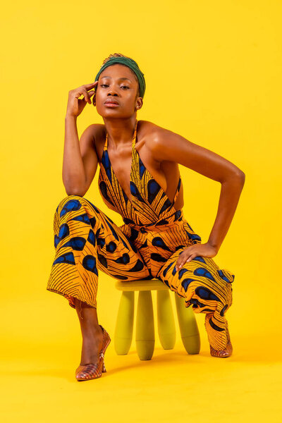 Young african woman with traditional costume on yellow background, sitting looking at camera