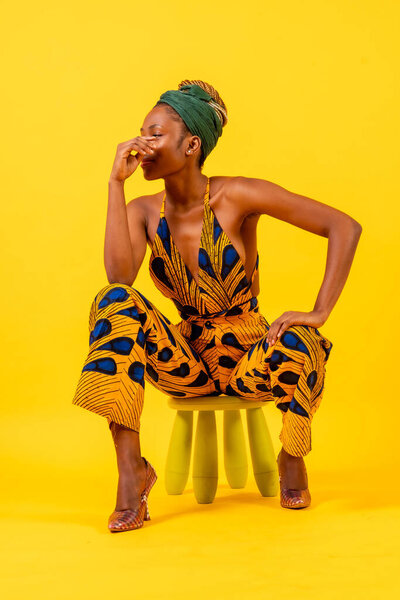 Young African woman in traditional dress on yellow background, sitting, commercial photography