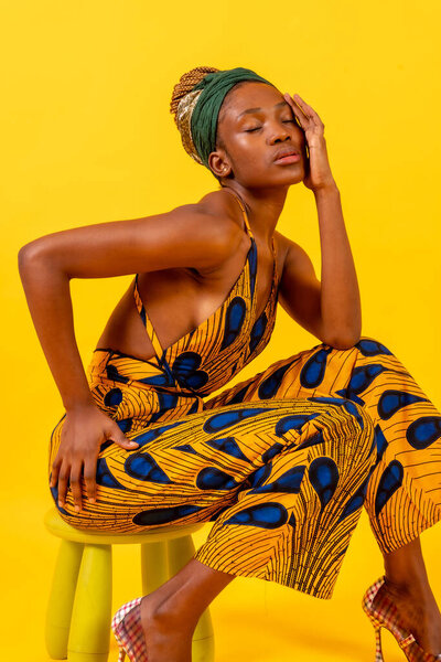 A person woman of black ethnicity with traditional costume in yellow background, sitting in a fashion pose