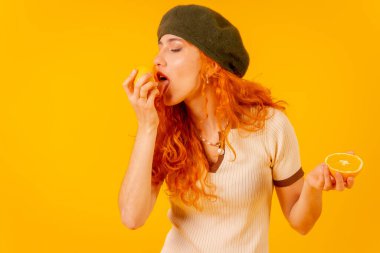 Kızıl saçlı kadın portakalı sarı arka planda tutuyor, meyveleri yiyor.