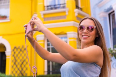 Sarışın, sarı ve mavi dış görünüşlü genç bir kadın telefonla fotoğraf çekiyor.