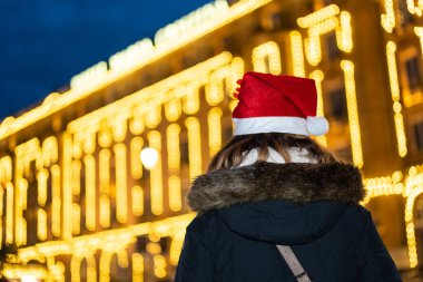 Arkadan bir kadın, gece şehrinde Noel şapkalı, aydınlanmış bir bina.