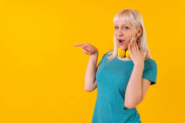 Pointing ads left with headphones, blonde caucasian girl on yellow background studio