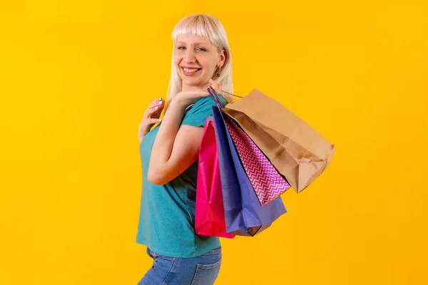 Laughing shopping with bags on sale, blonde caucasian girl in studio on yellow background