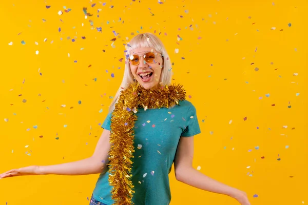 smiling party with confetti at a celebration, blonde caucasian girl in studio on yellow background