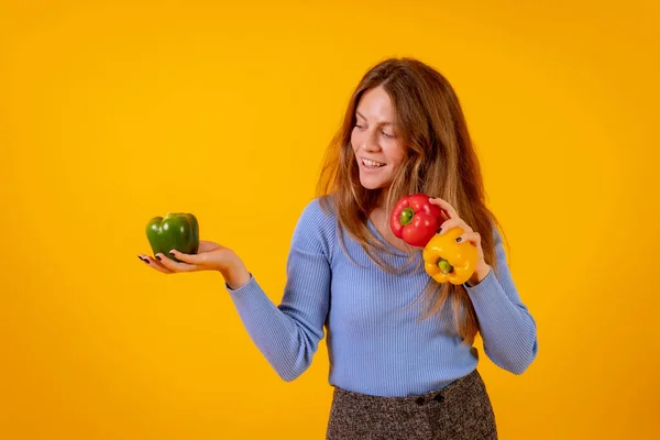 Vegan woman smiling with green, yellow and red bell peppers on a yellow background, vegetarian life