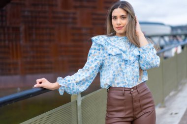 Young woman on a city river bridge smiling, lifestyle concept, blue shirt and brown pants