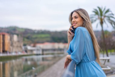 Blonde woman in blue dress smiling talking on the phone at the river in the city, lifestyle concept