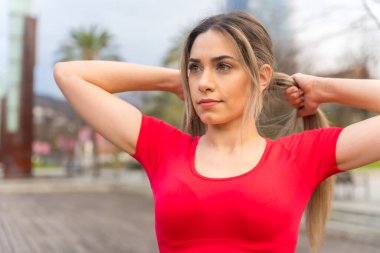 Young fit woman in red clothes doing sports in the city in spring, fitness, putting on ponytail