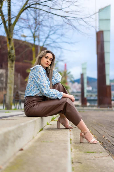 Young woman sitting in a park in the city, lifestyle concept, blue shirt and brown pants