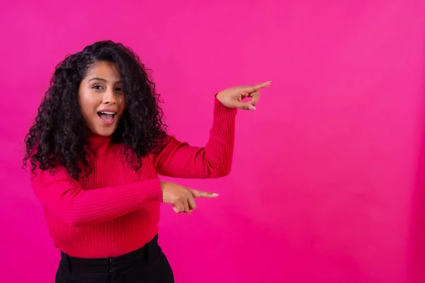 Curly-haired woman pointing to the right on a pink background, studio shot, copy paste