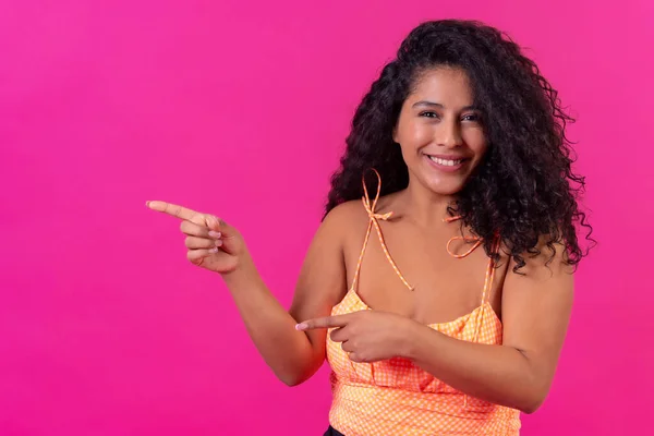 Curly-haired woman in summer clothes on a pink background pointing left, studio shot