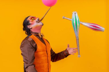 Portrait of a juggler in a vest with a painted face juggling with maces on a yellow background