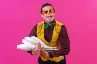 Juggler in a vest and with painted face juggling clubs on a pink background, cradling like a baby
