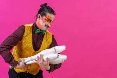 Juggler in a vest and with painted face juggling clubs on a pink background, cradling like a baby