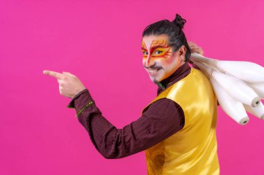 Juggler in a vest and with painted face juggling maces on a pink background, pointing