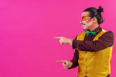 Clown with white facial makeup showing empty space from pink background, announcing something, pointing left