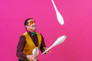 Clown with white facial makeup on a pink background, juggling maces